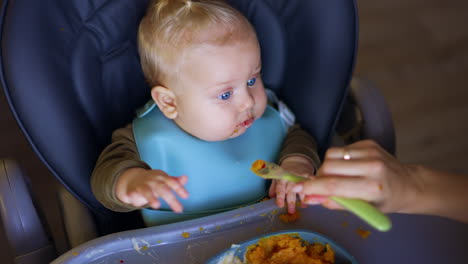 caucasian baby with adorable plump cheeks and blond hair sits in high chair. mother's hand giving spoon with pureed veggies to her child. top view.