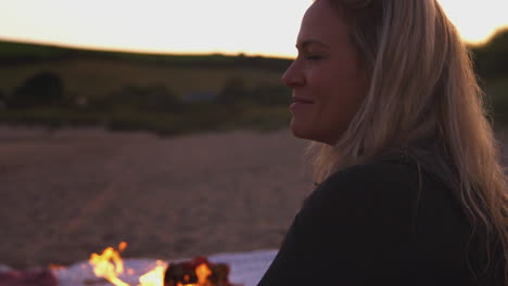 woman sitting by camp fire on beach as sun sets behind her