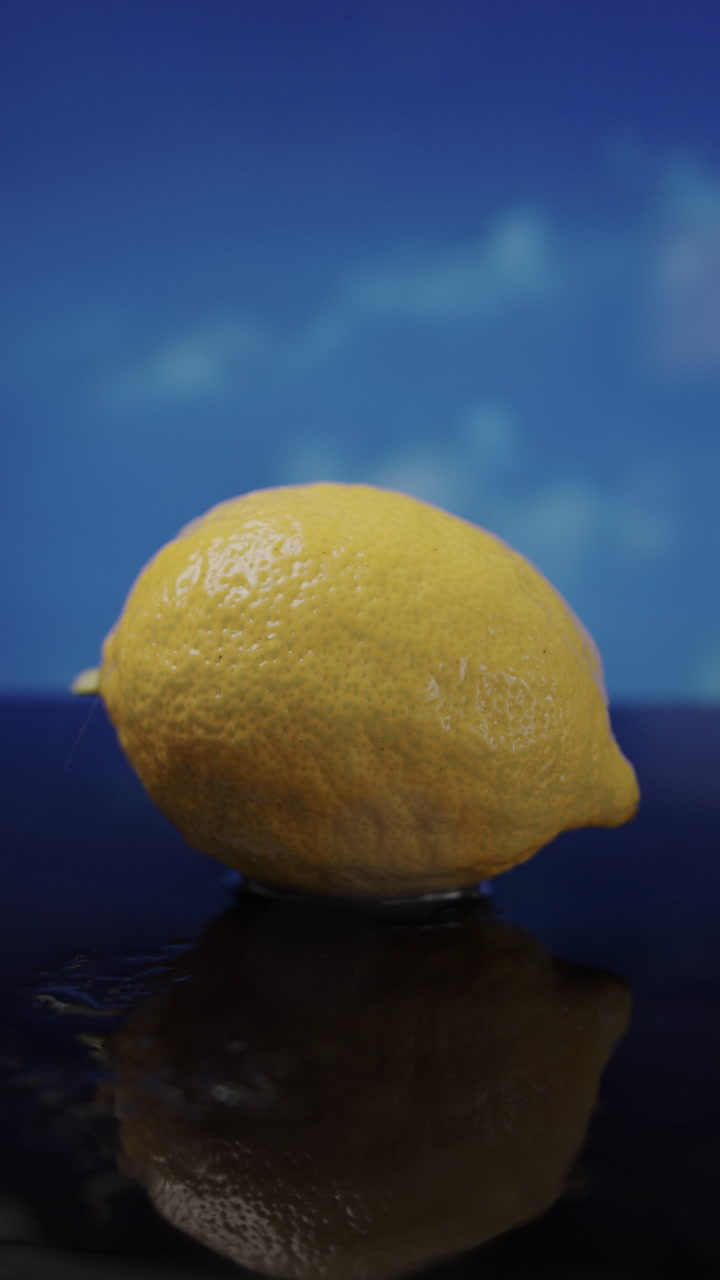 Vertical 9:16 video of a bright yellow lemon on a wet surface as raindrops fall and blue clouds move behind, creating strong color contrast