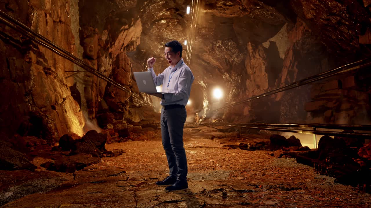 Full Body Side View Of An Asian Male Professional Worker Standing With His Laptop In Underground Mine Tunnel, He Raises His Fist Up With Screaming Goal After Typed On The Laptop