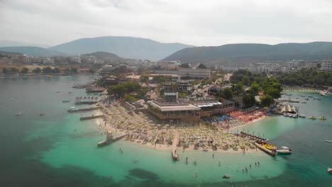 Luftaufnahme-Des-Wunderschönen-Ksamil-Strandes-Mit-Stadt-Im-Hintergrund