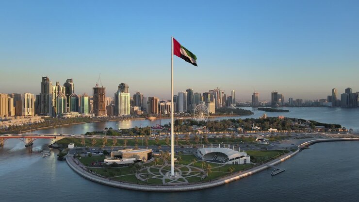 Aerial view of the Flag of the United Arab Emirates waving in the air, the Blue sky in Background, The national symbol of UAE over Sharjah's Flag Island, United Arab Emirates, 4K Video