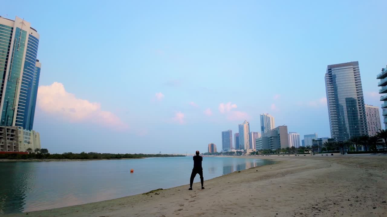 A man gracefully practices Tai Chi on an urban beach at sunrise in slow motion. The serene scene features tall city buildings, calm waters, and golden light illuminating the sky