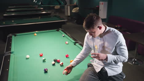 up shot of gentleman in white shirt and grey trousers sits on green pool table, sipping lemon drink with black straw in dimly lit billiard room. billiard balls scattered on table