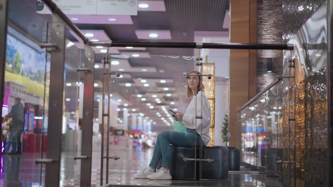 dama sentada en un moderno centro comercial visto a través del vidrio, con un ambiente animado con iluminación festiva y otros compradores en el fondo