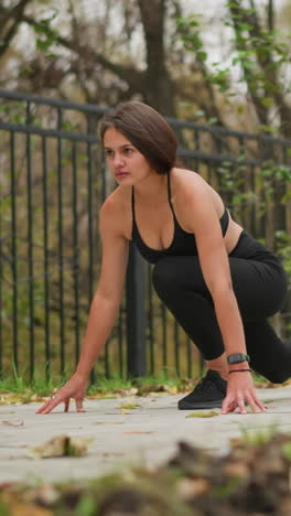 a woman squats down, focused and ready to run, dressed in black activewear, the background features an iron fence and dry trees, giving a sense of outdoor training and preparation