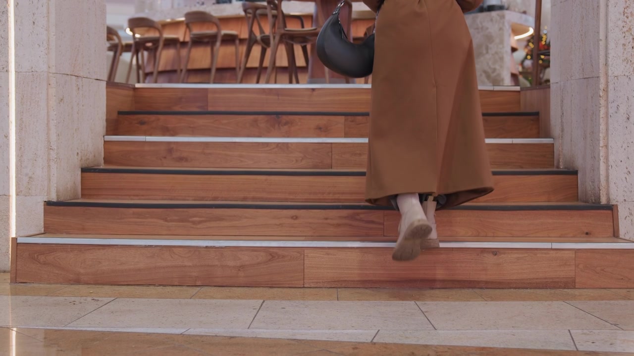 Woman ascending wooden stairs into cafe, long brown coat trailing, small round bag swinging, bare ankles visible on warm wooden steps, polished marble floor below, chairs and festive decor at landing