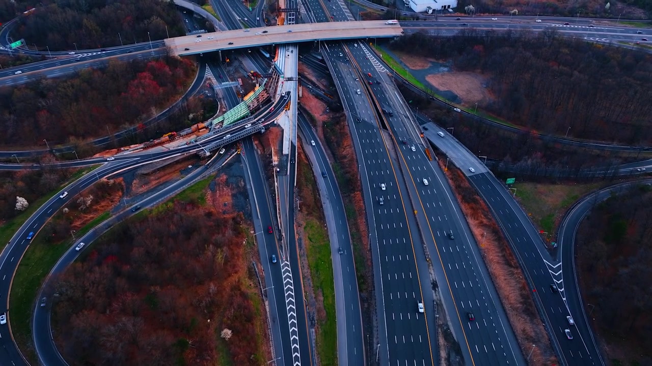 Aerial of a complex highway interchange with bridge work, New Jersey. Overhead of merging ramps and a flyover beside ongoing construction with warehouses and woodland around