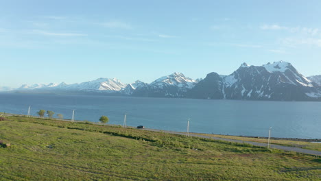 Aerial-view-following-a-car-driving-on-the-coast-of-the-arctic-ocean,-snowy-mountains-in-the-background,-sunny,-summer-day,-in-Spaakenes,-Nordland,-Norway---low,-dolly,-drone-shot