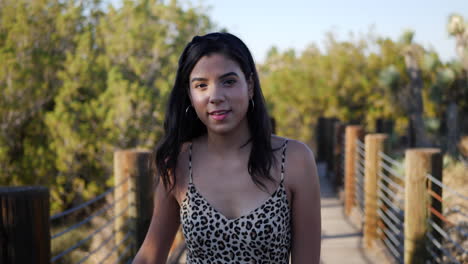 Cute-hispanic-woman-walking-in-slow-motion-along-a-wooden-bridge-outdoors-in-sunlight-smiling-and-laughing