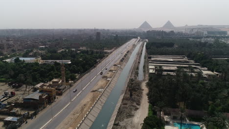 Toma-Aérea-Rastreando-Y-Elevándose-Hacia-Las-Pirámides-De-Egipto-En-Giza-En-El-Fondo-De-Un-Brazo-Del-Río-Nilo-En-Primer-Plano-Rama-Maryotya-Que-Muestra-Las-Tierras-Agrícolas-Y-La-Vegetación