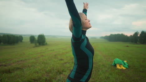 una mujer en un campo de hierba al aire libre practica yoga, levantando y bajando lentamente su mano con un enfoque tranquilo, mientras que un par de botas amarillas y una tela verde se ven en el fondo de la hierba archivada.