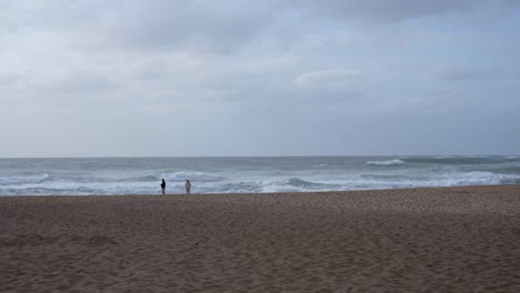 Tourists-walking-on-a-beach-with-violent-sea-waves-crashing-at-Praia-da-Adraga-in-Portugal