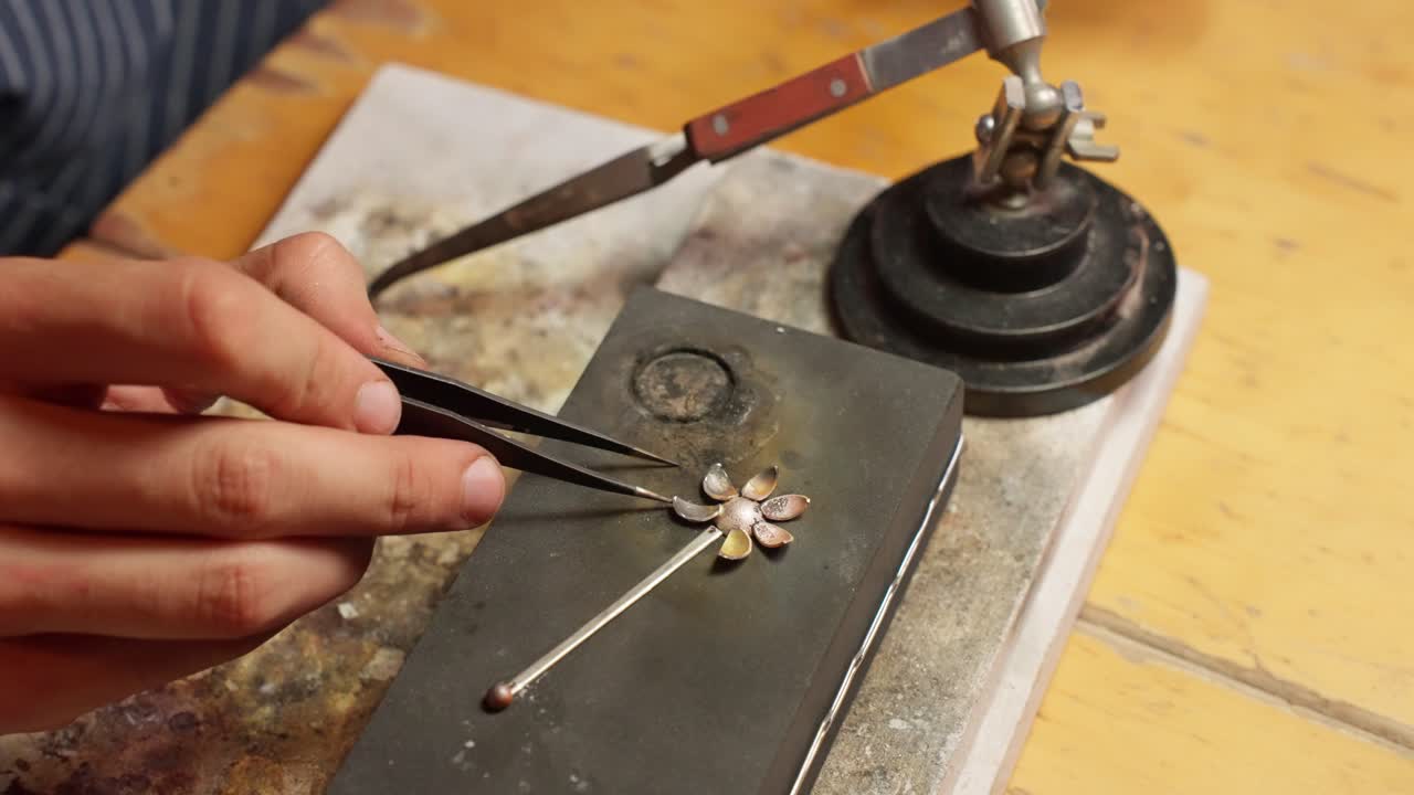 An artisan delicately creates a silver flower pendant with tweezers and metalworking tools on a wooden workbench.