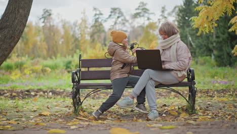 woman seated on bench working on laptop as her child rushes from behind holding bright yellow autumn leaf to share discovery in leafy park surrounded by golden foliage and scattered leaves
