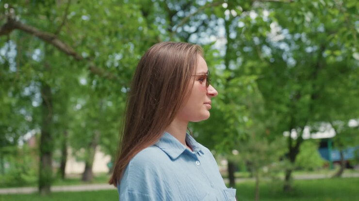 Donna serena con occhiali da sole che si gode un ambiente tranquillo sotto un cielo soleggiato mentre si crogiola alla luce del sole estivo all'aperto, donna rilassata con occhiali da sole che si gode il sole estivo in uno scenario pittoresco del parco
