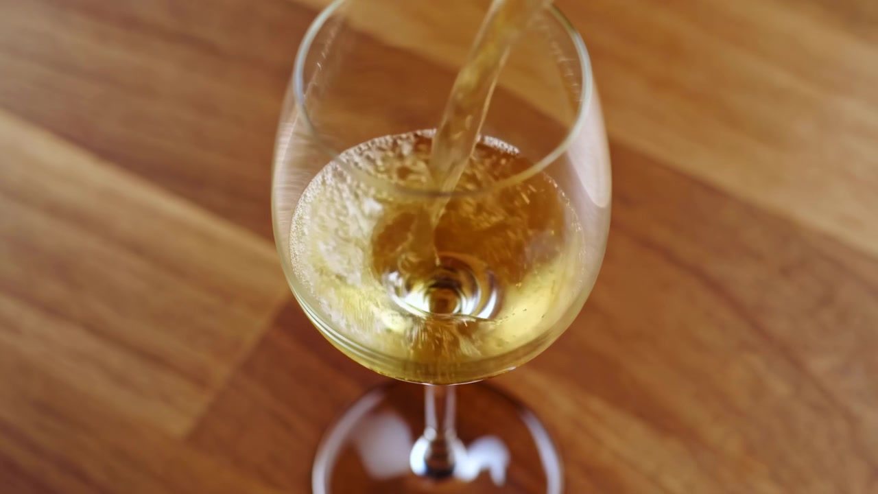 Une scène sereine et élégante de vin blanc versé progressivement dans un verre sur une surface de table en bois, capturée d'une perspective de haut en bas. Ce moment visuellement attrayant est parfait.