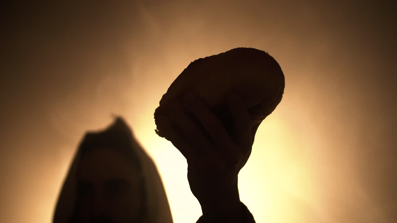 Silhouette of Jesus Christ in white hooded robe holding bread as he stands in glowing, bright light praying. Perfect for representation of bread of life, last supper, Easter Christianity visual.