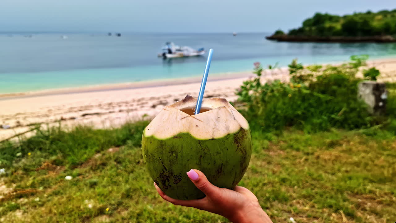 Hand holds green coconut with straw, preparing to sip fresh juice on tropical beach afternoon