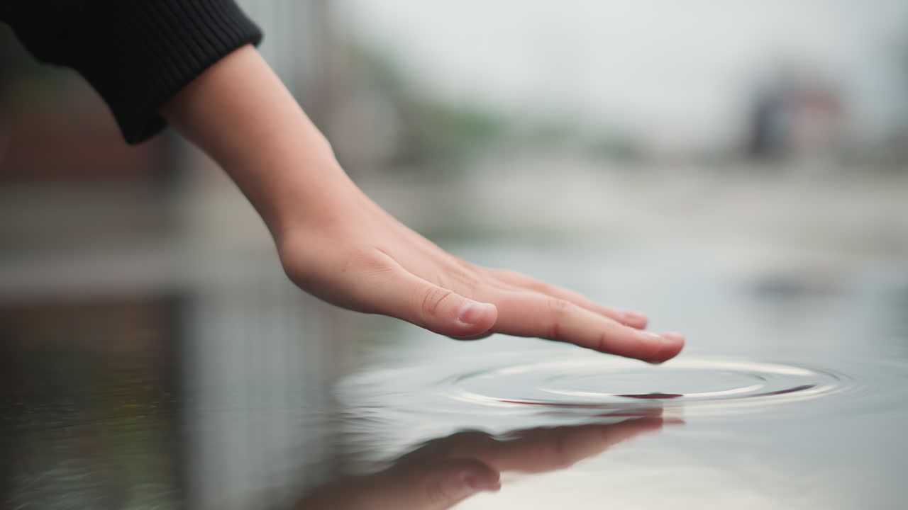 Mano di donna bianca che tocca una pozzanghera, delicato polpastrello che crea increspature su una superficie urbana bagnata, luce soffusa e nuvolosa, primo piano della manica, umore riflessivo, curiosità nostalgica, rallentatore.