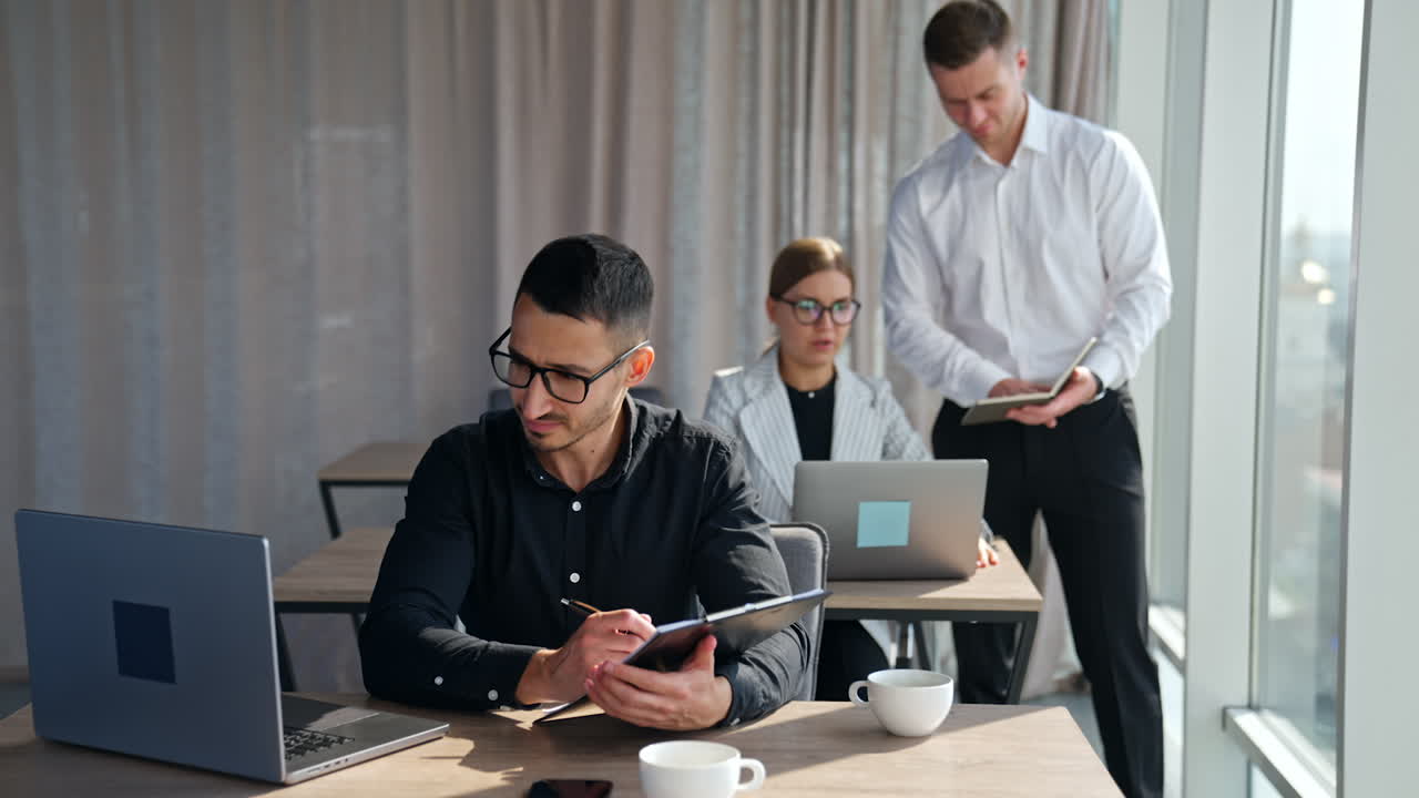 Male manager standing at the desk of female employee checking the data in his notebook. Male brunet colleague focused on the screen of his laptop.
