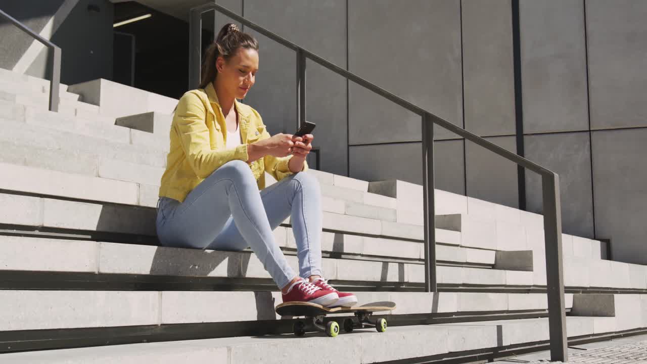 Caucasian woman sitting on stairs with skateboard, using smartphone on sunny day