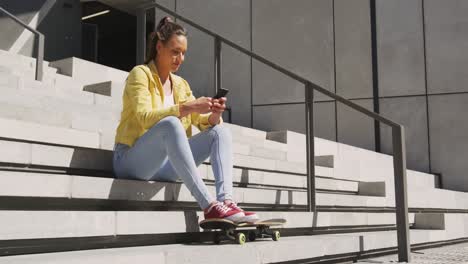 Caucasian-woman-sitting-on-stairs-with-skateboard,-using-smartphone-on-sunny-day