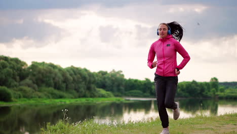 a girl in a pink jacket and black pants runs near the river in headphones preparing for the marathon