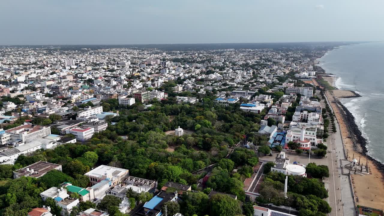 This aerial stock footage presents a wide, high-angle view of a coastal city and its beachfront. A sprawling city extends inland, anchored by a large, lush green park in the center