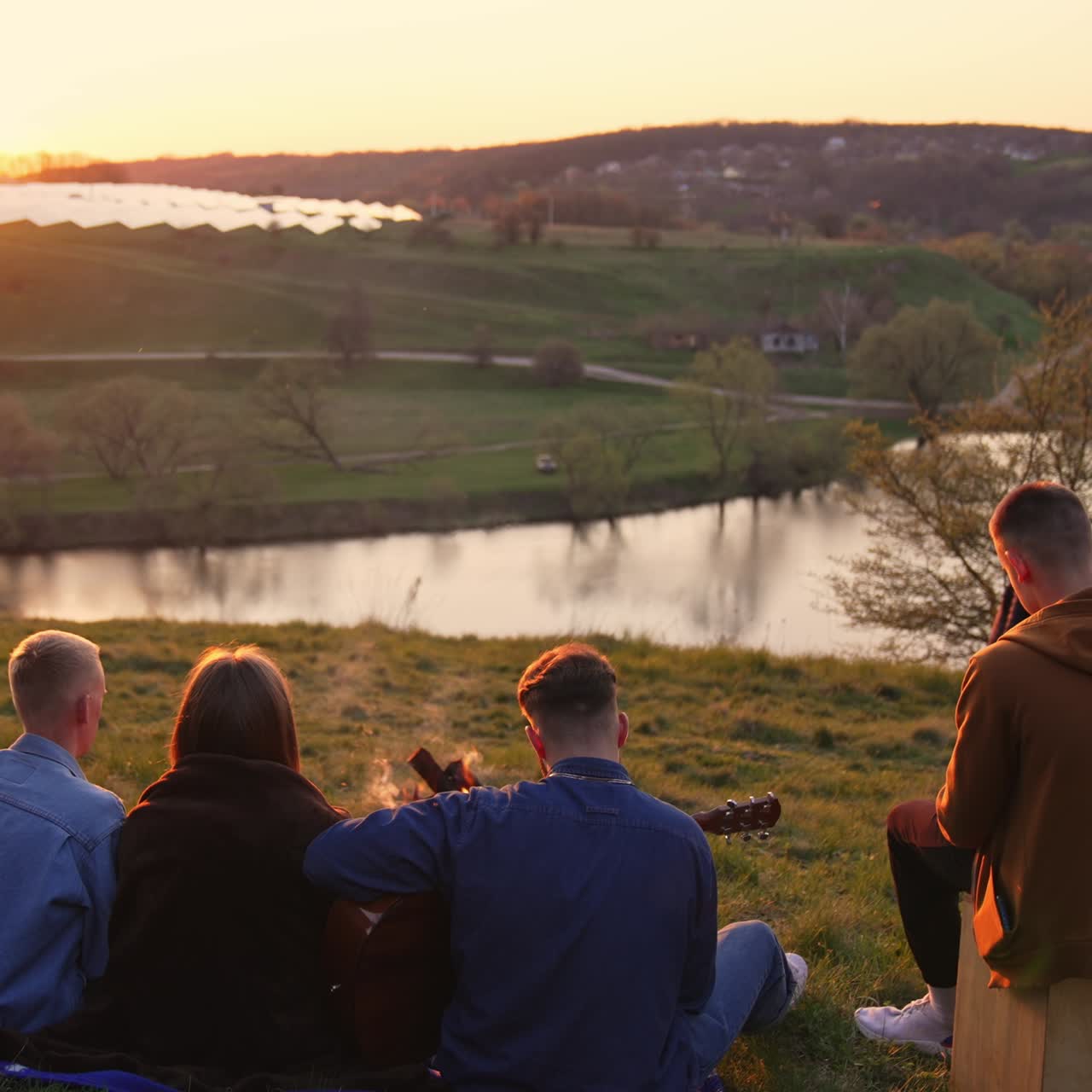 Group of friends sitting on the slope of hill and playing musical instruments. Young company enjoying sunset and summer evening