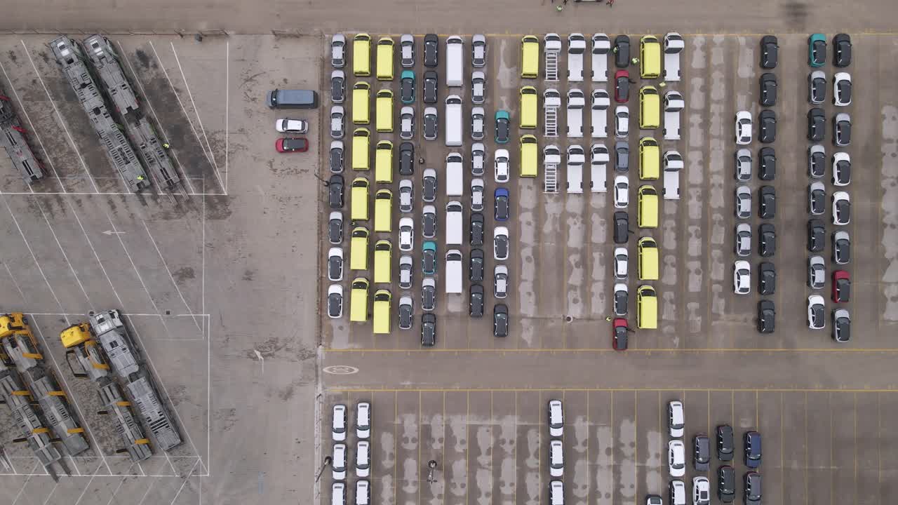 An aerial perspective showcasing a large parking lot with rows of vehicles, including cars, yellow vans, and transport trucks. The vehicles are organized in sections.