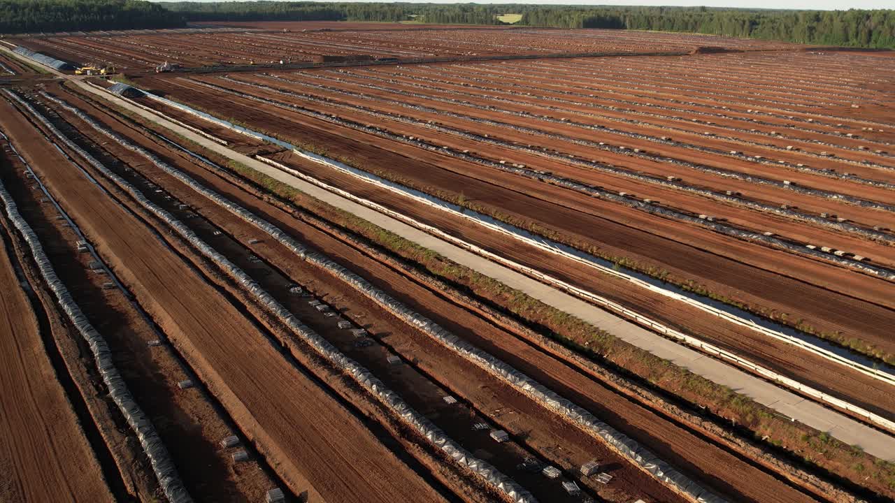 Aerial view of a large peat bog harvesting site with long parallel rows of extracted peat, wooden pallets, and covered stacks stretching across the landscape