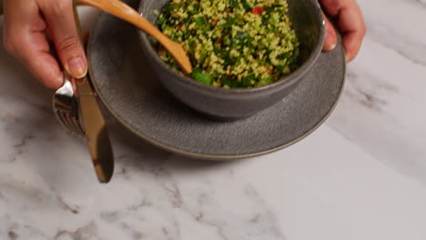 close up of woman at home in kitchen preparing healthy vegetarian or vegan meal eating bowl of tabbouleh salad