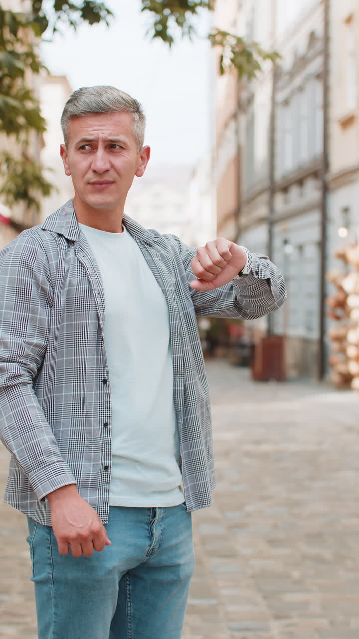 Mature caucasian man tourist with serious face looking at watch while walking on street in city