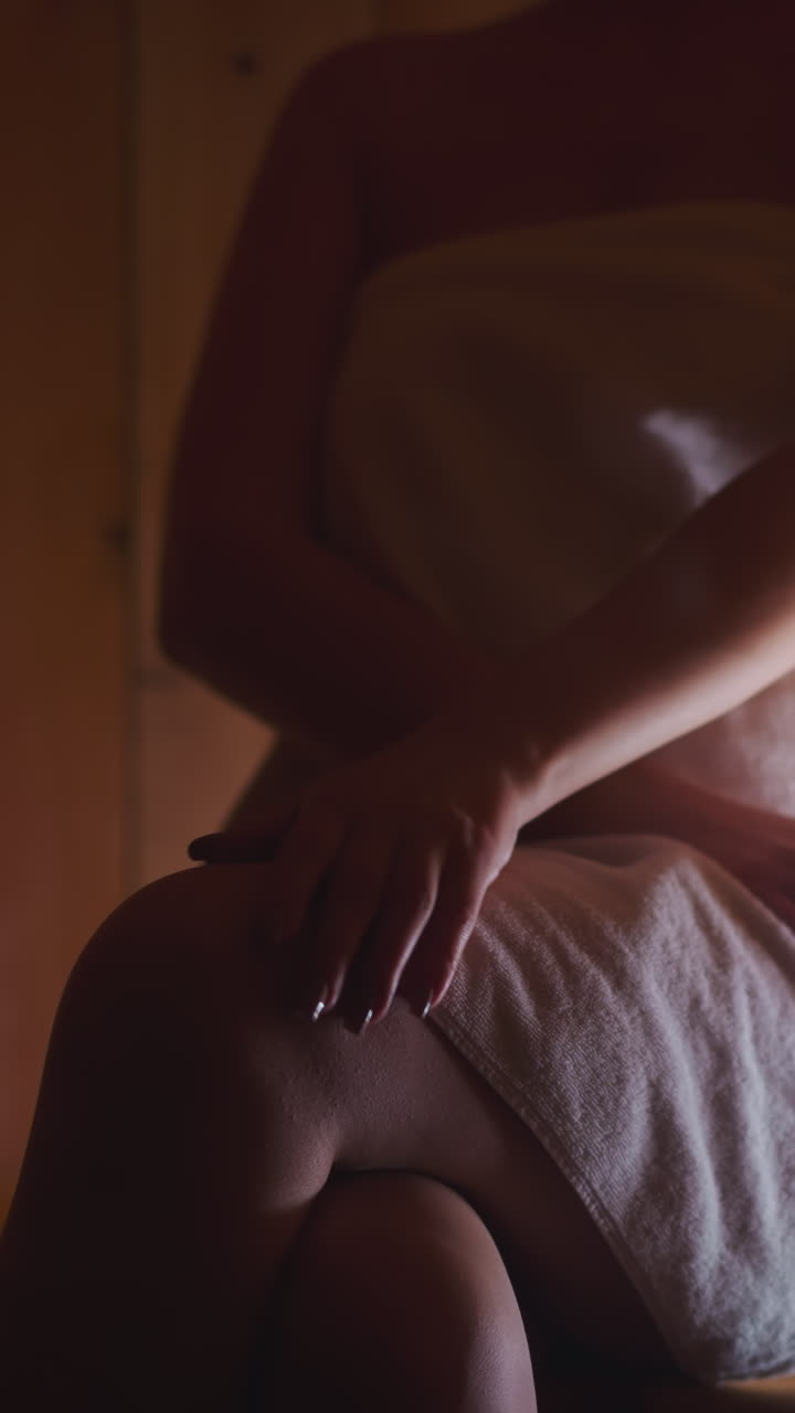 Woman strokes knee while sitting in sauna. Young woman shows stamina sitting in hot semi-dark sauna in towel. Wellness center spa procedures
