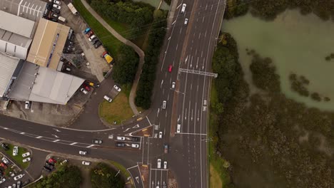 traffic scene on junction of highway in auckland, nz. cloudy summer day at sunset. aerial top down. driving cars turning into road. grass fields and bush and lake at takapuna area