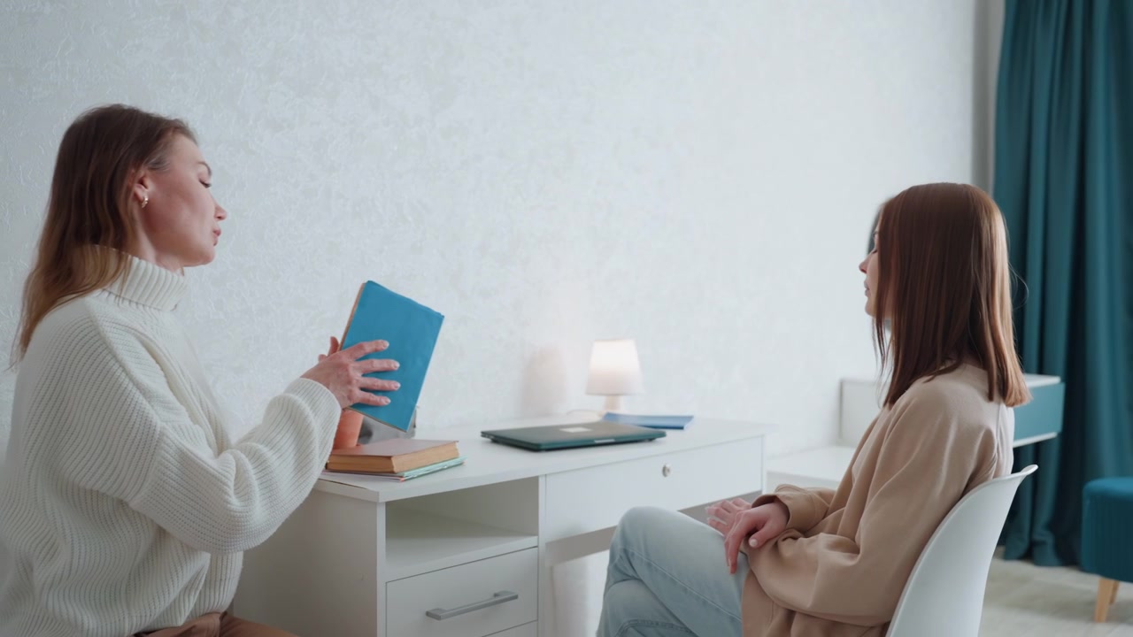 Intens feedbacksession med hvid coach, to kvinder i lyst soveværelseskontor, stående og siddende billeder, lærer viser blå mappe, peger på noter, studerende lytter, fokuseret øjenkontakt, fast udtryk