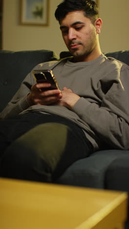 vertical video of young man spending evening at home sitting on sofa with mobile phone scrolling through internet or social media