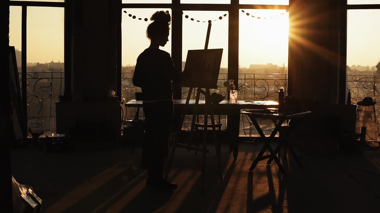A figure of female artist shaded by sunset on background drawing on easel. Inspirational video.