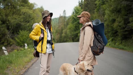 Two-girls,-a-blonde-and-a-brunette,-in-special-tourist-hiking-clothes-with-large-backpacks,-together-with-their-light-colored-dog,-stand-near-the-road-and-communicate-against-the-backdrop-of-a-green-forest
