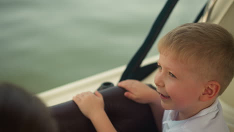 delighted little boy enjoys motorboat sailing on summer day closeup. smiling child travels by tourist boat along calm sea water. summertime entertainment