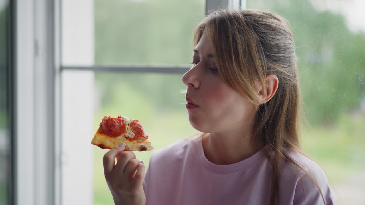 Student on holiday enjoying grilled egg pizza sitting peacefully close to window wearing casual purple t-shirt, gazing outside thoughtfully with natural green blurred background
