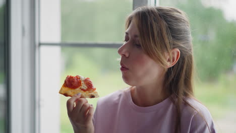 student on holiday enjoying grilled egg pizza sitting peacefully close to window wearing casual purple t-shirt, gazing outside thoughtfully with natural green blurred background