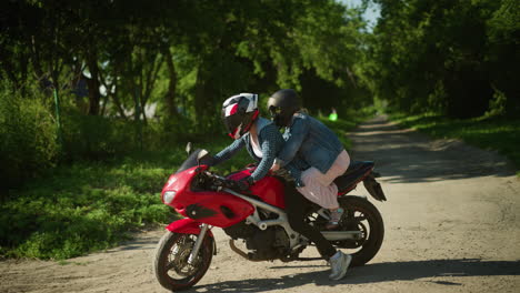 two sisters ride a red power bike, both wearing helmets, as they drive into a paved walking path surrounded by trees, a blur view of buildings can be seen in the background