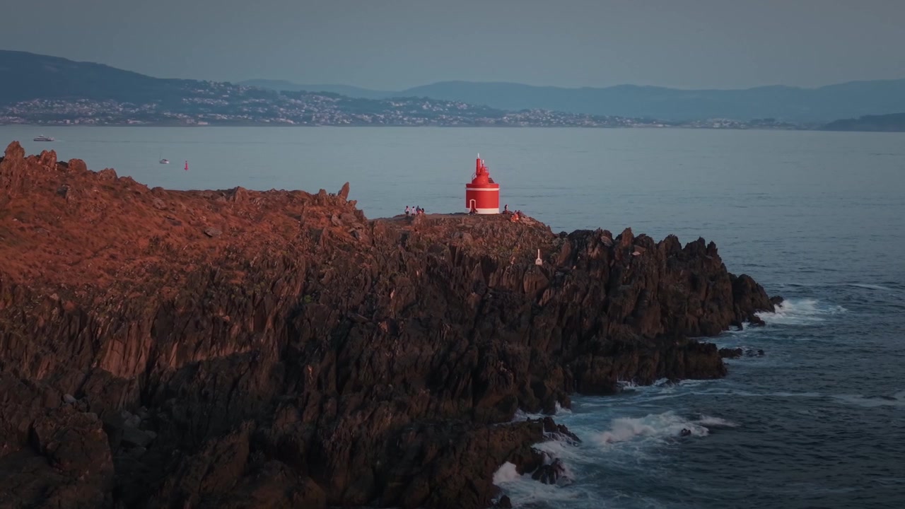 Ripresa aerea in orbita del faro di Punta Robaleira su scogliere rocciose in Galizia, Spagna, al tramonto con le calme acque dell'Atlantico