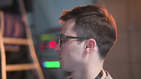 person in tv studio, viewed from side, focused on adjusting video equipment. wearing glasses and working with control panel, studio lighting and equipment visible in background