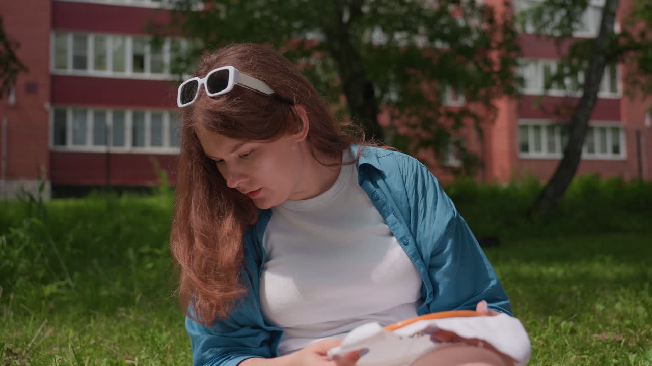 Girl wearing blue shirt seated outdoors adjusting hair while holding embroidered fabric in hand under bright sunlight, surrounded by lush greenery and red building in soft background