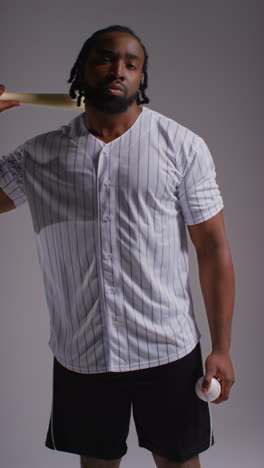 Vertical-Video-Studio-Portrait-Of-Serious-Male-Baseball-Player-Wearing-Team-Shirt-Holding-Bat-Turning-Around-Shot-Against-Grey-Background