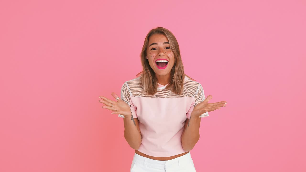 dama rubia con manos tatuadas, vestida de arriba. ella está mirando sorprendida y diciendo wow, sonriendo mientras posa contra un fondo rosa. cerca