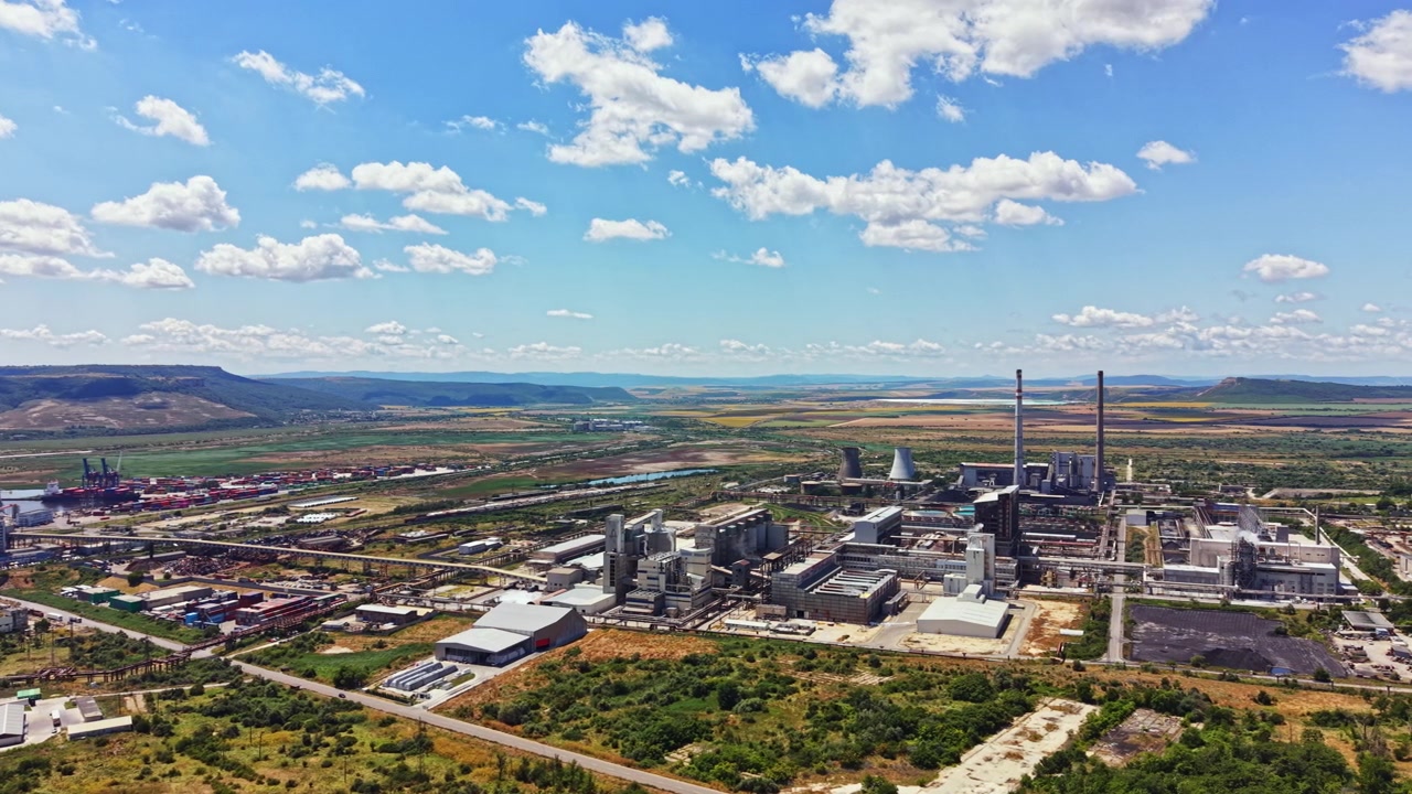 Zona industrial muestra fábricas, chimeneas y contenedores cerca de un río con colinas y nubes al fondo durante el día